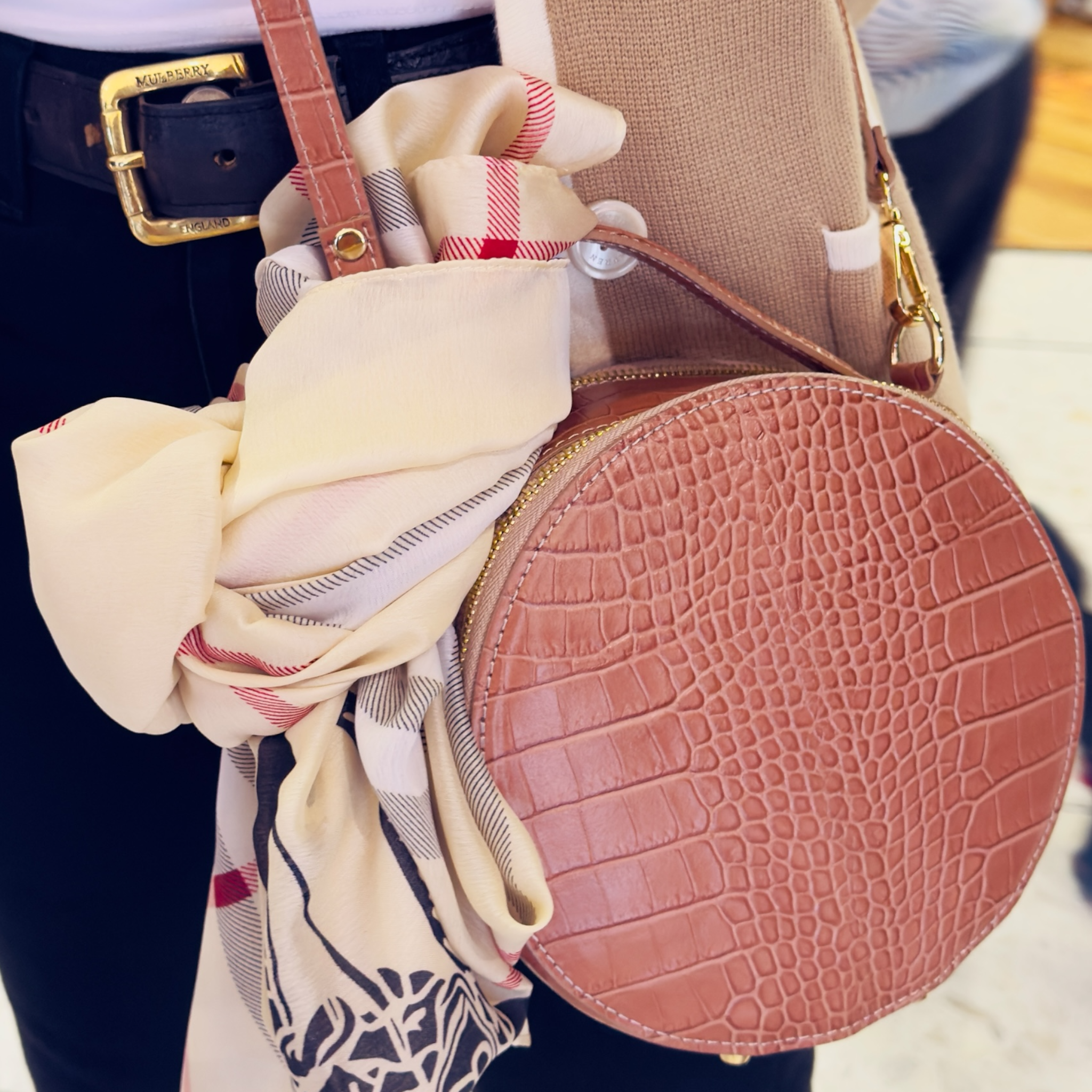 A lady wearing a round bag across her body with a scarf tied on it. 
