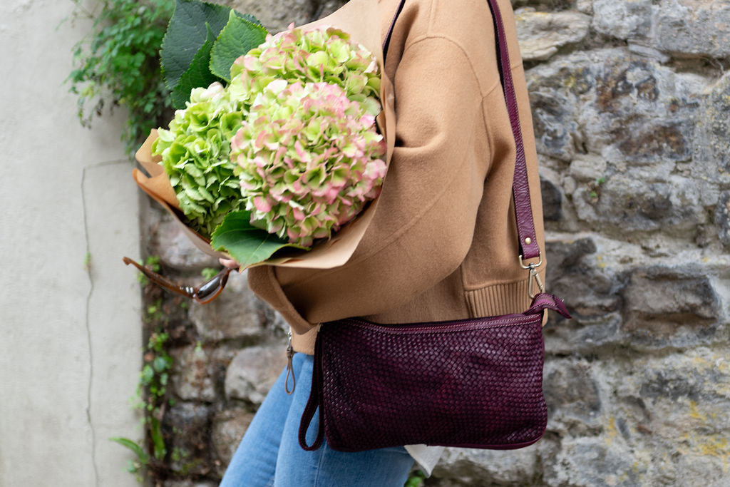 Italian handwoven leather pouch colour Aubergine 
Removable leather shoulder / wrist strap 
Imported from Florence 
Sussex Kent Surrey Independent handbag retailer
Person holding a pouch with flowers  